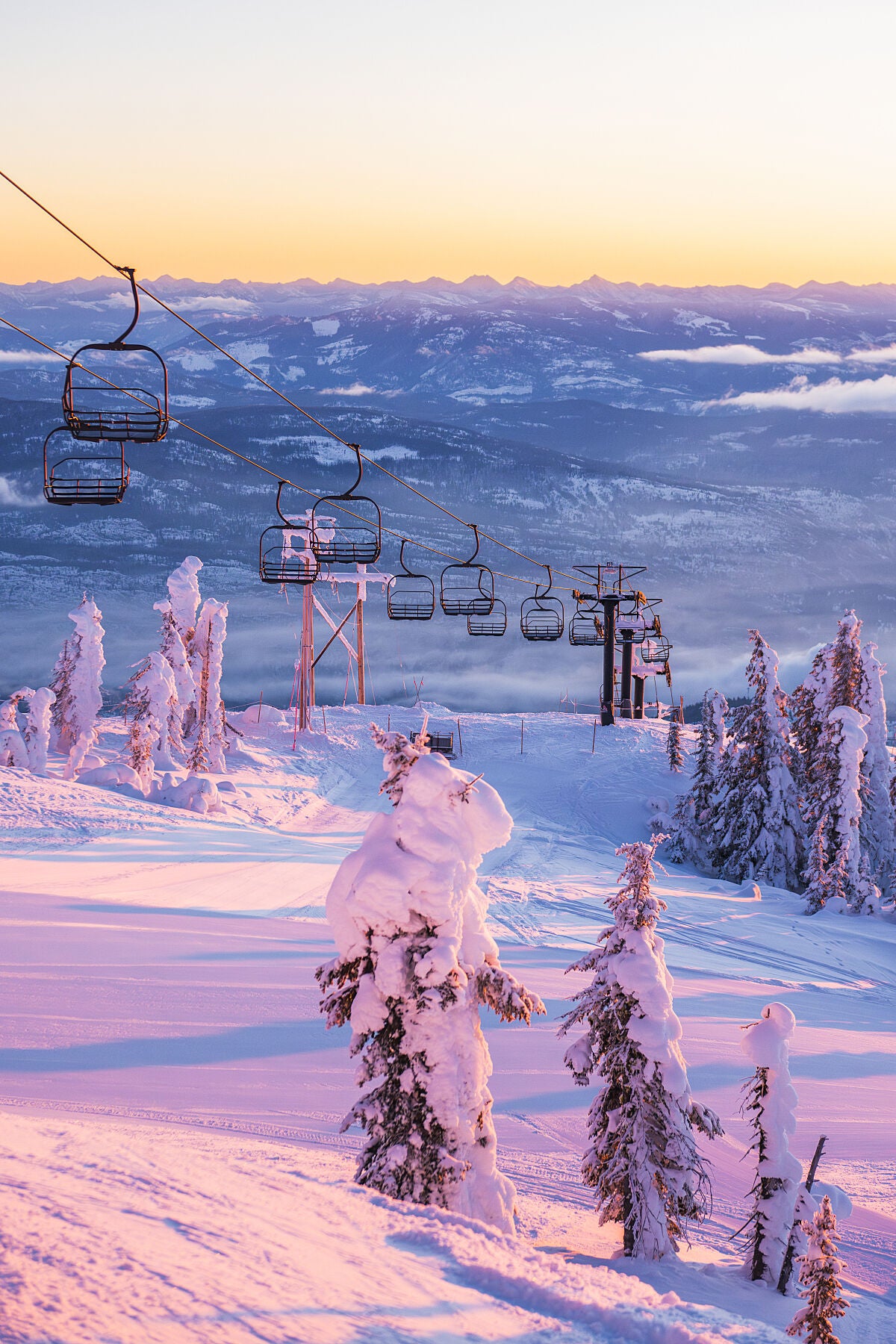 A photograph featuring the Motherlode chairlift at Red Resort with a pastel sunrise light on the slopes of Granite Mountain. The chairlift is in the foreground, and the background is a gradient of pink to purple hues with snow-covered mountains, fine art landscape print by Ashley Voykin