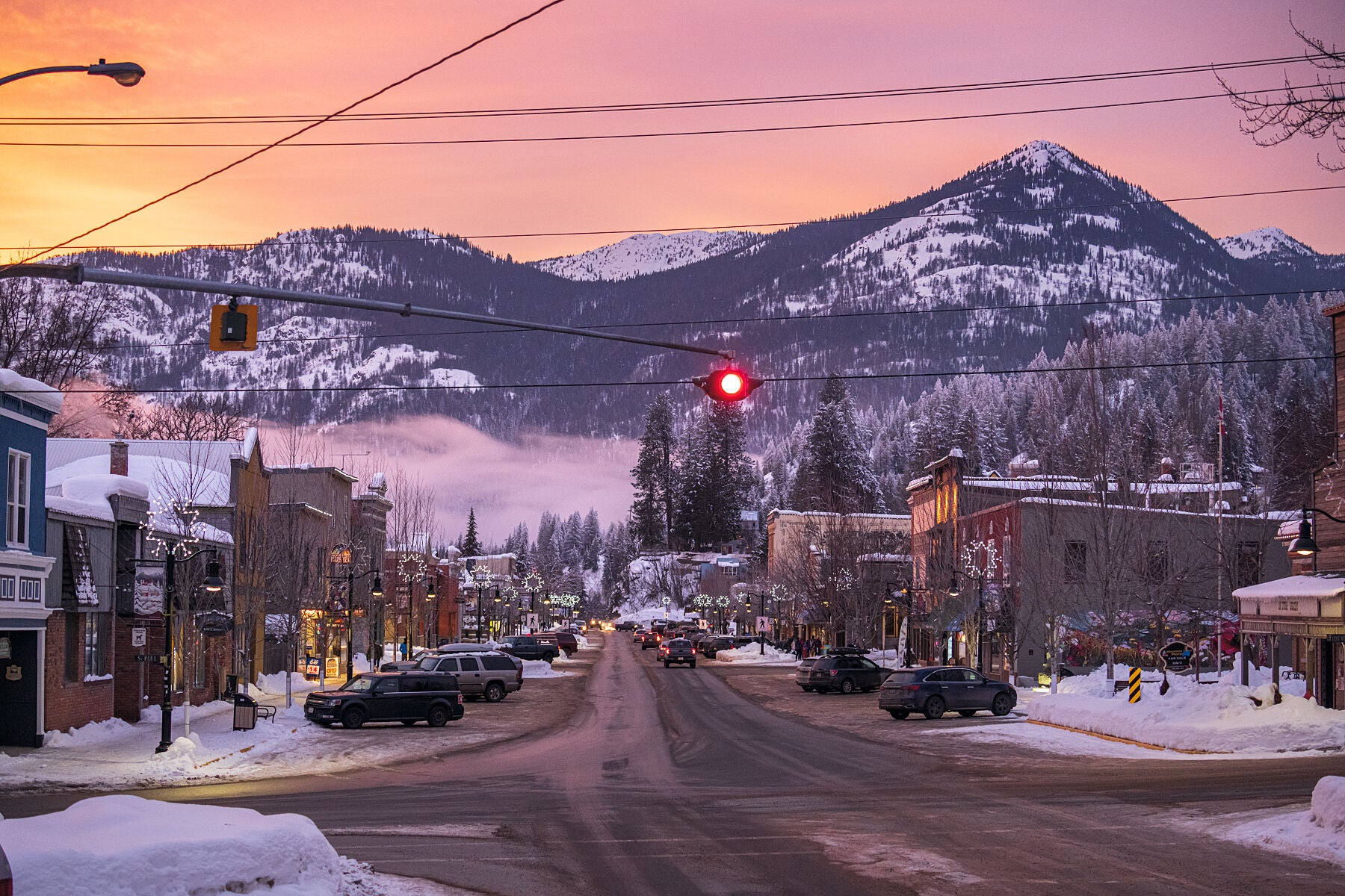 A winter sunset in Rossland, British Columbia, with a red traffic light at the intersection of two streets, surrounded by snow-covered buildings and mountains in the distance. Fine art landscape photograph by Ashley Voykin.