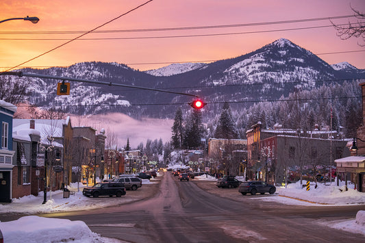 A winter sunset in Rossland, British Columbia, with a red traffic light at the intersection of two streets, surrounded by snow-covered buildings and mountains in the distance. Fine art landscape photograph by Ashley Voykin.