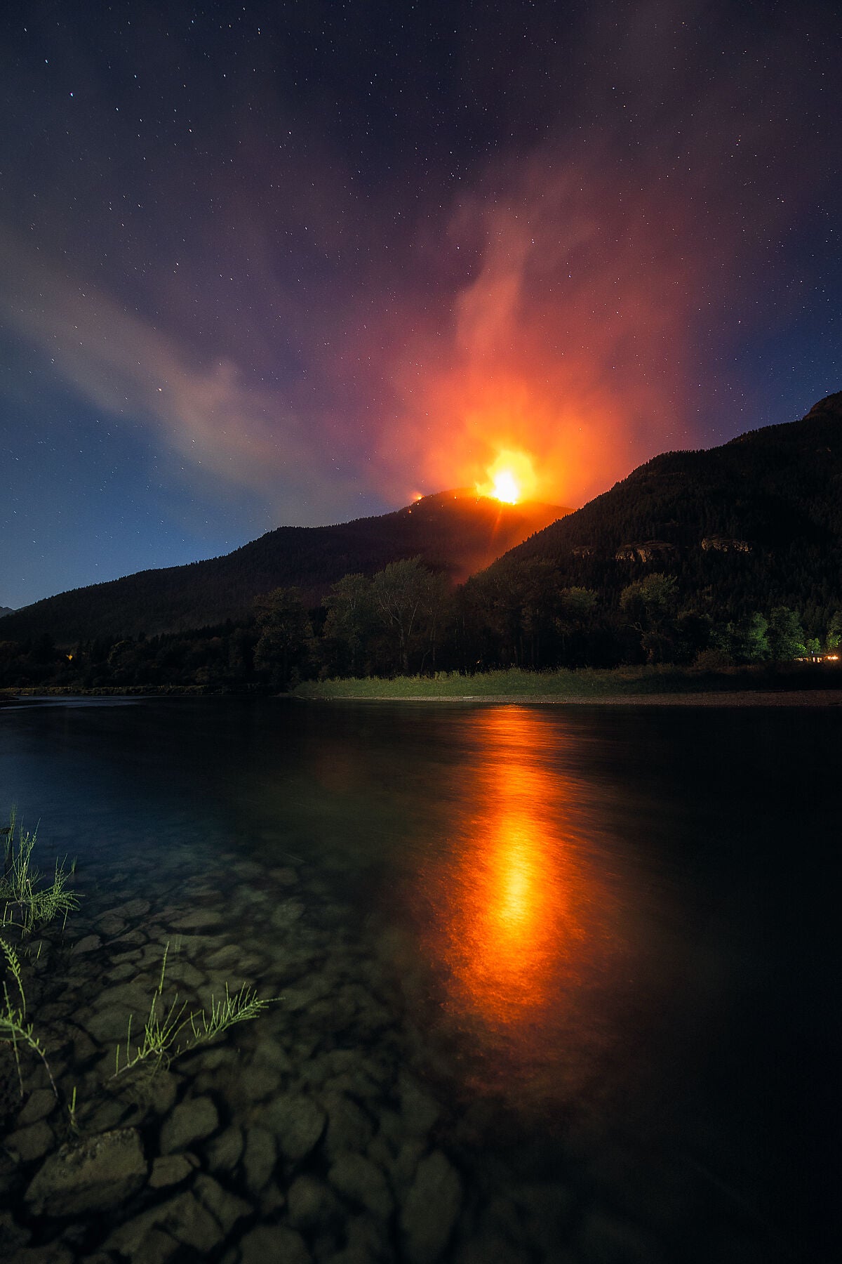 A photograph featuring the Talbot Creek wildfire above the Slocan River, with a vibrant red glow from the fire reflecting on the water's surface and a dark sky in the background. Photography by Ashley Voykin.