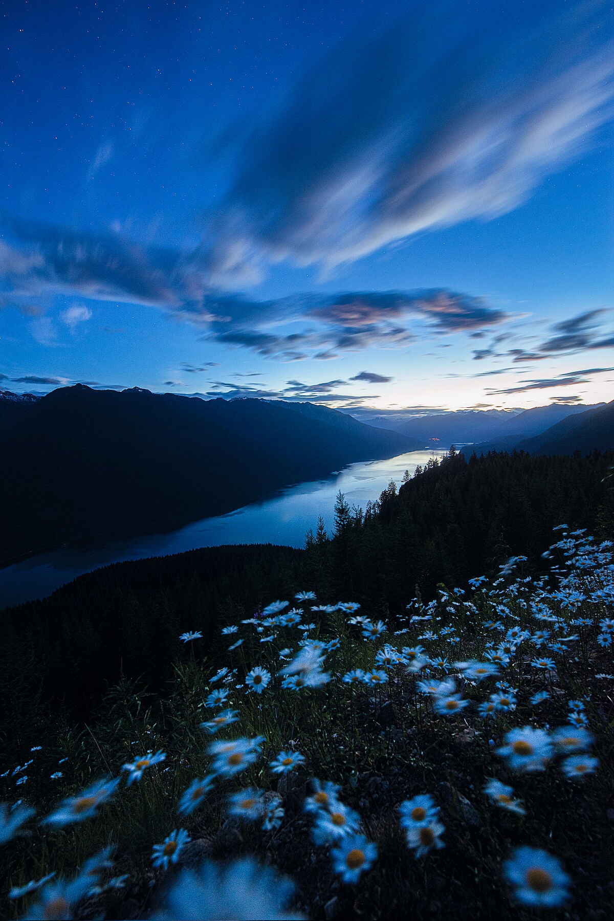A landscape photo featuring a serene view of Slocan Lake at dusk with mountains in the background and a clear night sky filled with stars. Fine Art Photography by Ashley Voykin.
